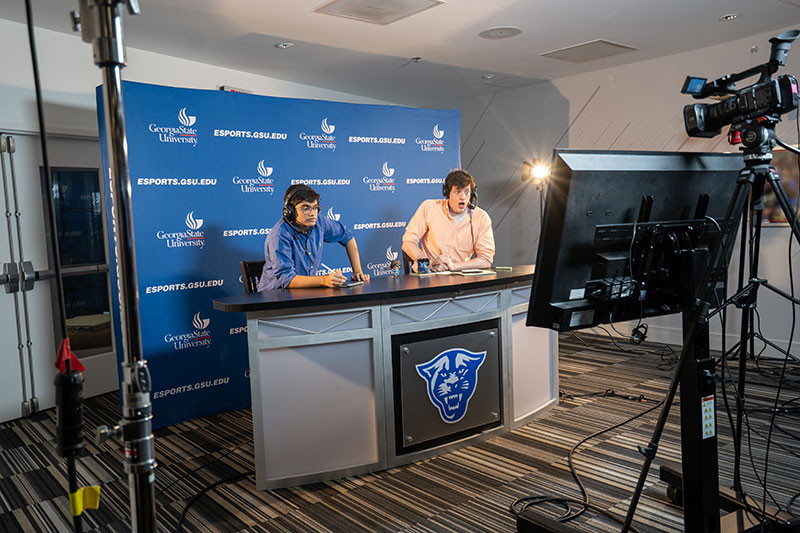 2 Shoutcasters behind a counter. An Esports.GSU.EDU backdrop is behind them and a camera in front of them.