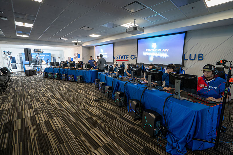 Two long tables . Each table has 6 monitors on top and 6 computers in front hooked to the monitors. A competitor sits in front of each monitor.