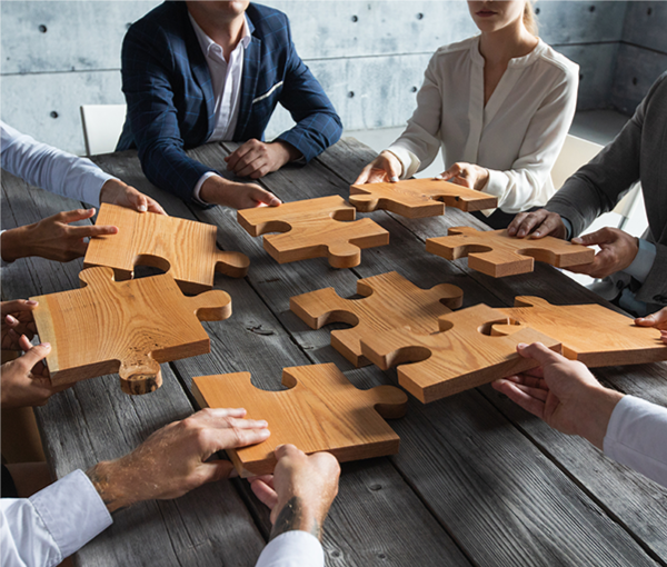 people around a table. Each person is holding a large puzzle piece and they are trying to fit them together.